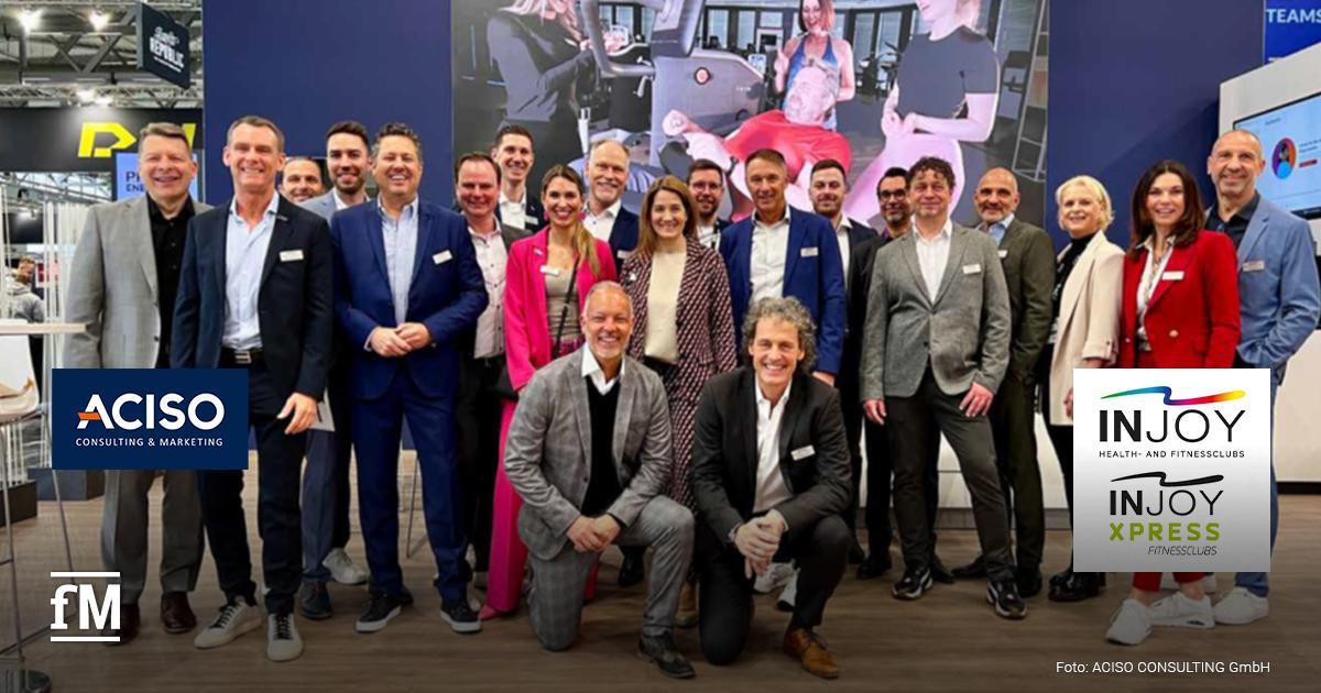 Eine große Gruppe von Menschen steht auf der FIBO 2025 vor einem Messestand von ACISO Consulting & Marketing sowie INJOY Health- und Fitnessclubs. Die Teilnehmer tragen elegante Business-Outfits, viele mit Namensschildern, und lächeln in die Kamera. Im Hintergrund ist eine große Leinwand mit einem Bild aus einem Fitnessstudio zu sehen. Das ACISO-Logo ist links im Bild platziert, das INJOY-Logo rechts unten. Das fM-Logo befindet sich in der unteren linken Ecke.