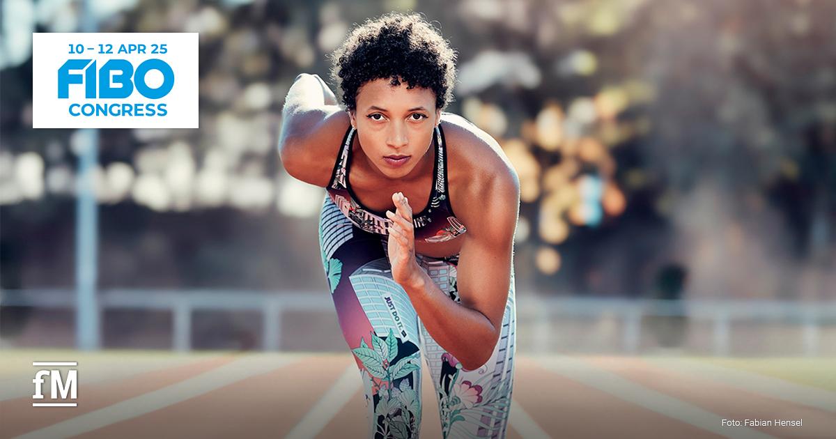 Olympiasiegerin im Weitsprung Malaika Mihambo blickt in Startpose auf der Tartanbahn eines Stadions direkt in die Kamera. Im Hintergrund sind Büsche und davor ein weißer Zaun und eine Hochsprunganlage zu erahnen.