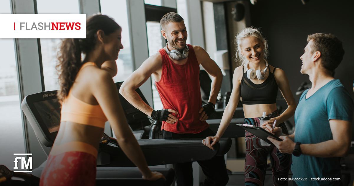 Eine Gruppe junger, sportlicher Menschen unterhält sich gut gelaunt in einem modernen Fitnessstudio. Zwei Frauen und ein Mann stehen in Sportkleidung vor Laufbändern, während ein Trainer mit einem Klemmbrett in der Hand mit ihnen spricht. Alle wirken motiviert und lachen. Im oberen linken Bereich des Bildes befindet sich eine rote und schwarze Textbox mit der Aufschrift 'FLASH NEWS'. Unten links ist in weißer Schrift das Logo 'fM' platziert, das für die Marke 'fitness MANAGEMENT' steht. Die Bildquelle ist 'BGStock72 – stock.adobe.com'.