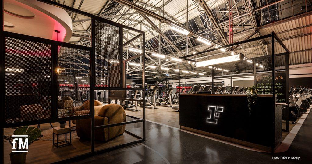 Ein modernes Fitnessstudio mit industriellem Design und offenen Metallstrukturen. Der Eingangsbereich ist mit einer stilvollen Rezeption ausgestattet, die ein beleuchtetes 'F'-Logo der Marke Fitness First zeigt. Im Hintergrund befinden sich zahlreiche Fitnessgeräte, darunter Laufbänder und Kraftmaschinen. Eine gemütliche Lounge mit Sesseln und einem kleinen Tisch ist durch ein Metallgitter abgetrennt. Die Beleuchtung ist warm und hebt die Architektur hervor