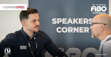 Zwei Männer im Gespräch beim FIBO Congress 2024, im Hintergrund das Schild „Speakers Corner“. Links trägt ein Mann ein Namensschild und dunkles Hemd. Rechts ein Mann mit Brille und Glatze. Links oben steht ein Gewinnspielhinweis. Rechts oben des FOBO Congress-Logo für 2025