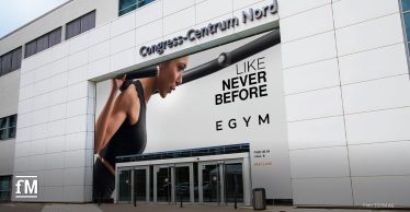 FIBO 2025 Eingang mit großflächiger EGYM-Werbung: Eine Athletin hebt eine Langhantel, begleitet vom Slogan 'Like Never Before'. Der Messestandort Congress-Centrum Nord ist zu sehen.