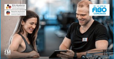 Ein lächelnder Trainer zeigt einer sportlich gekleideten Frau ein Tablet. Beide stehen in einem modernen Fitnessstudio. Logos der BSA-Akademie, DHfPG und des FIBO Congress 2025 sind zu sehen.