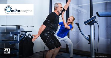 Ein älterer Mann trainiert mit EMS-Technologie von miha bodytec, während eine Trainerin anleitet. Er trägt einen EMS-Anzug. Die Trainerin in blauer Sportkleidung unterstützt ihn aktiv. Im Hintergrund ist ein modernes Studio mit Glaswänden zu sehen. Links oben befindet sich das miha bodytec-Logo, unten links das fM-Logo.