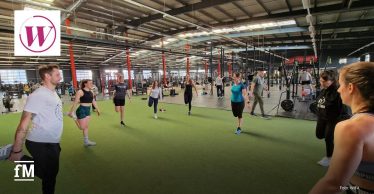 Eine Gruppe von Frauen und Männern nimmt an einem Warm-up in einem großen, modernen Fitnessstudio teil. Sie stehen auf einer grünen Trainingsfläche und führen Dehnübungen durch. Der Raum ist mit Kraftgeräten, Langhanteln und Trainingsstationen ausgestattet. Links oben im Bild befindet sich das Logo von WIFA, unten links das fM-Logo.